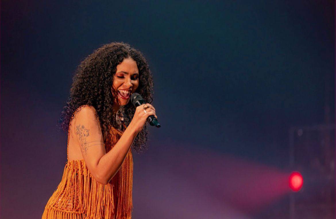 Mulher negra, com cabelos cacheados, longos e soltos, canta. Ela está posicionada na lateral esquerda da foto e virada em direção ao lado direito. A mulher, de batom vermelho e vestido de franjas marrom, segura um microfone.