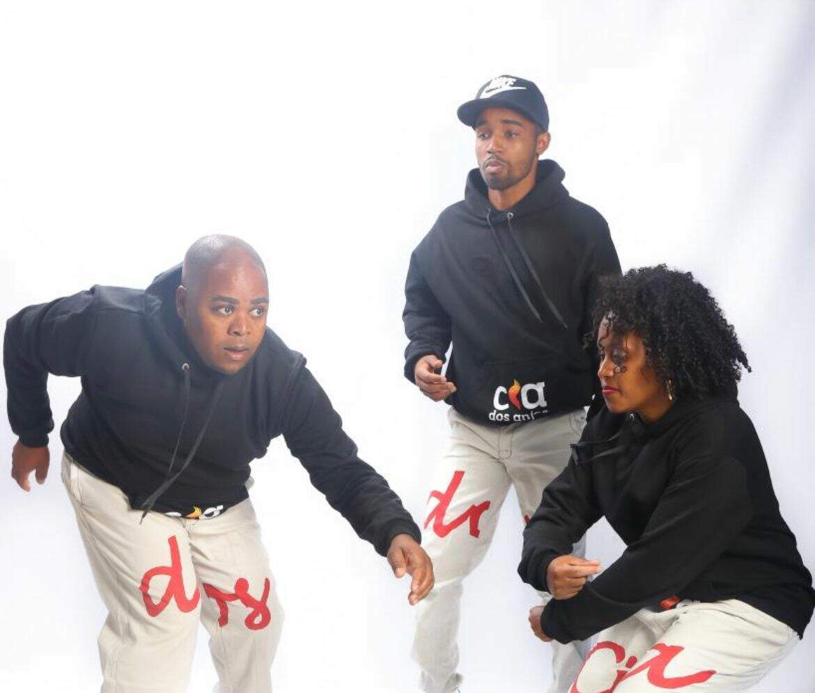 Em um fundo branco, dois homens e uma mulher aparecem em posição de dança. Os três são pessoas negras e estão usando roupas iguais: um moletom preto e uma calça branca