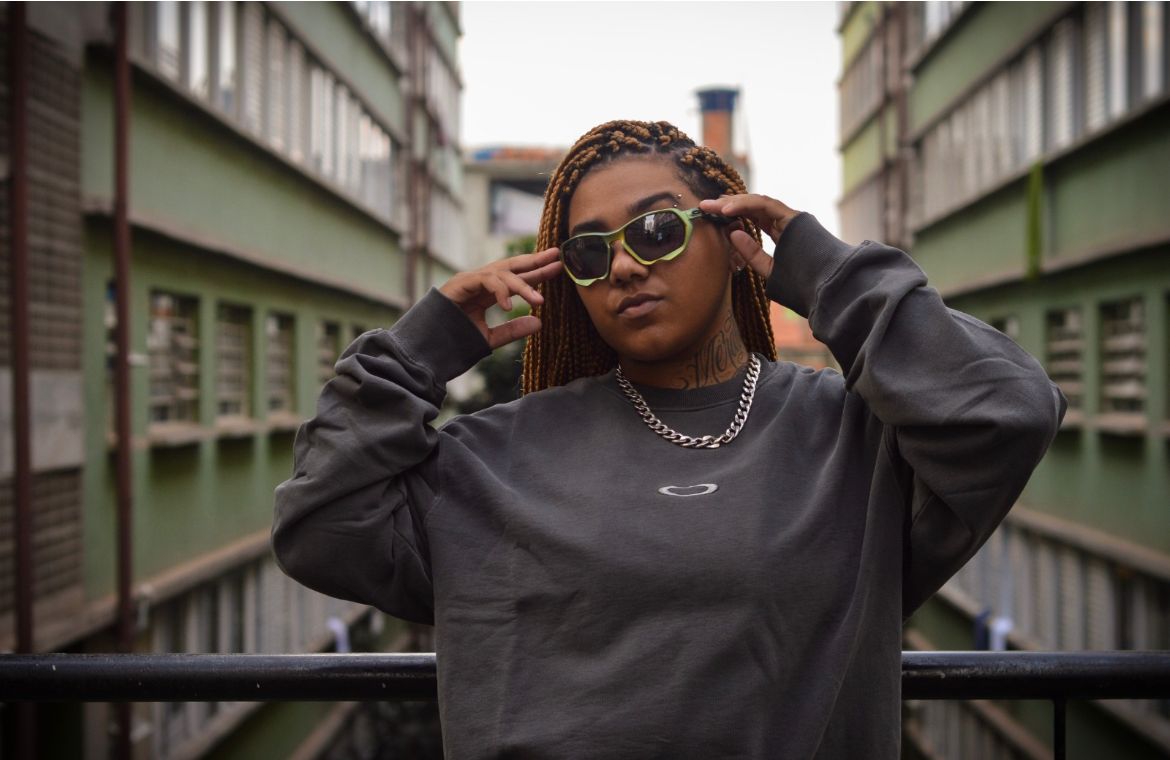 Mulher negra, com tranças no cabelo se posiciona diretamente para câmera. Ela está segurando um óculos escuro no rosto. A mulher usa um moletom cinza e corrente no pescoço.