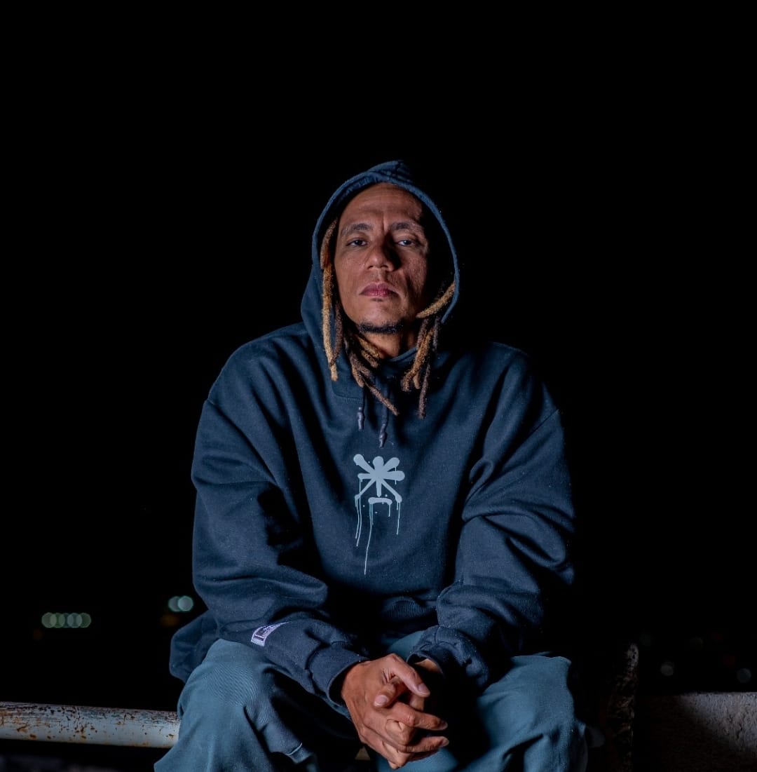 Homem negro com semblante sério olhando para câmera. Ele usa dreads loiros e um moletom com capuz azul. Esta sentado com as mãos juntas