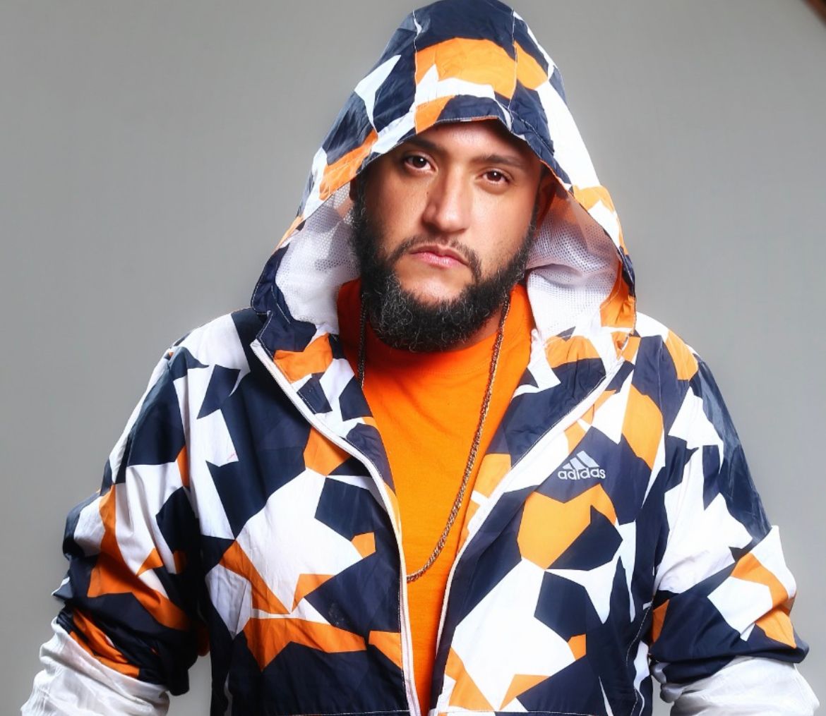 Homem negro, com barba, olhando diretamente para câmera. Ele está usando uma camisa laranja e um casaco com capuz colorido em tons laranja, azul e branco
