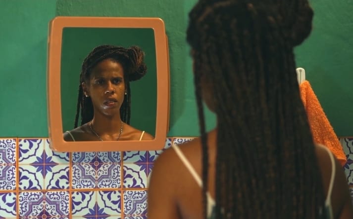 Mulher negra olhado para um espelho fixado na parede. Ela usa tranças. Na imagem só é possível ver o rosto dela pelo reflexo do espelho