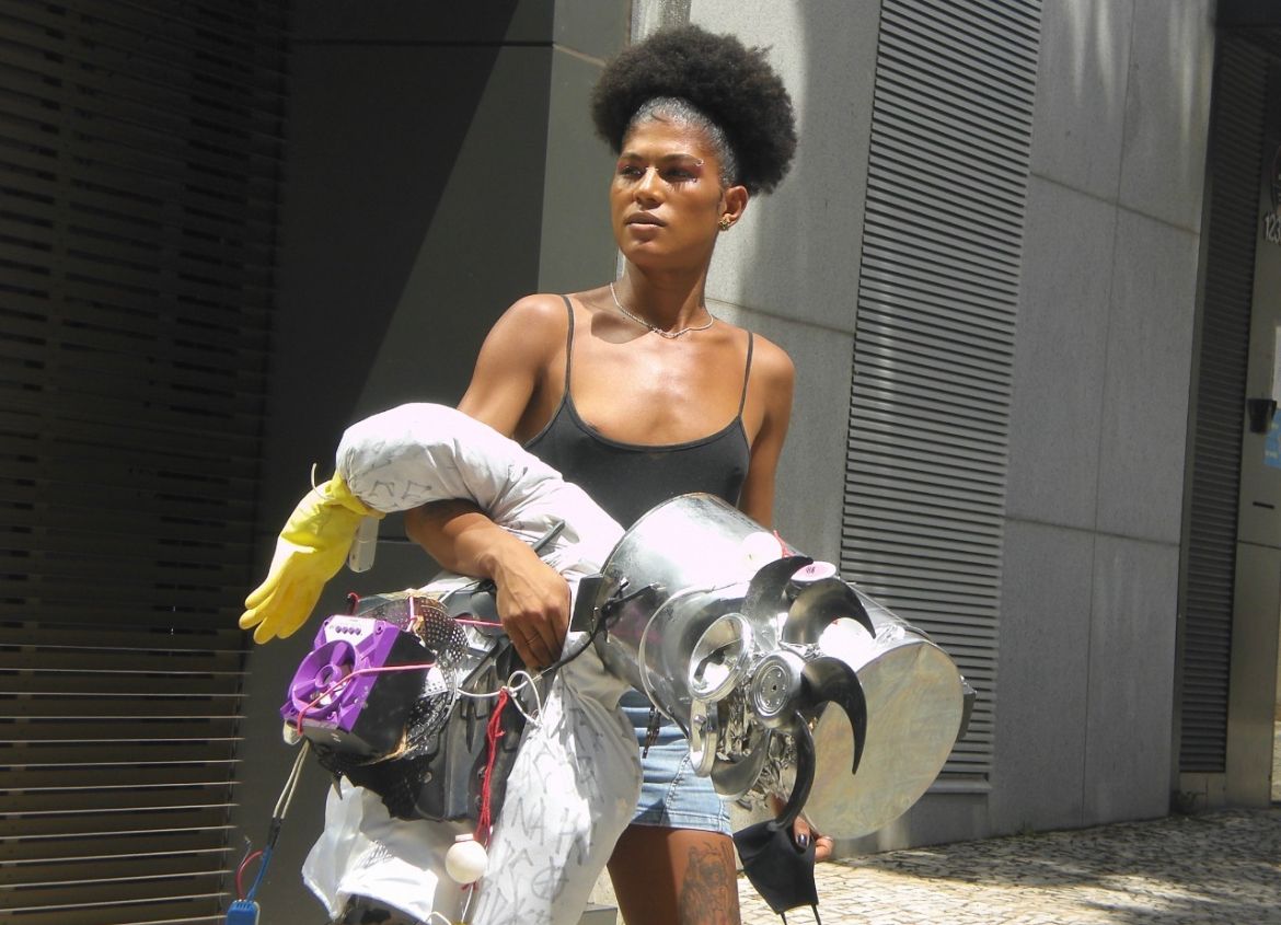 Mulher negra, de regata preta. Ela tem cabelos curtos, black power. A mulher está segurando adereços em metal.