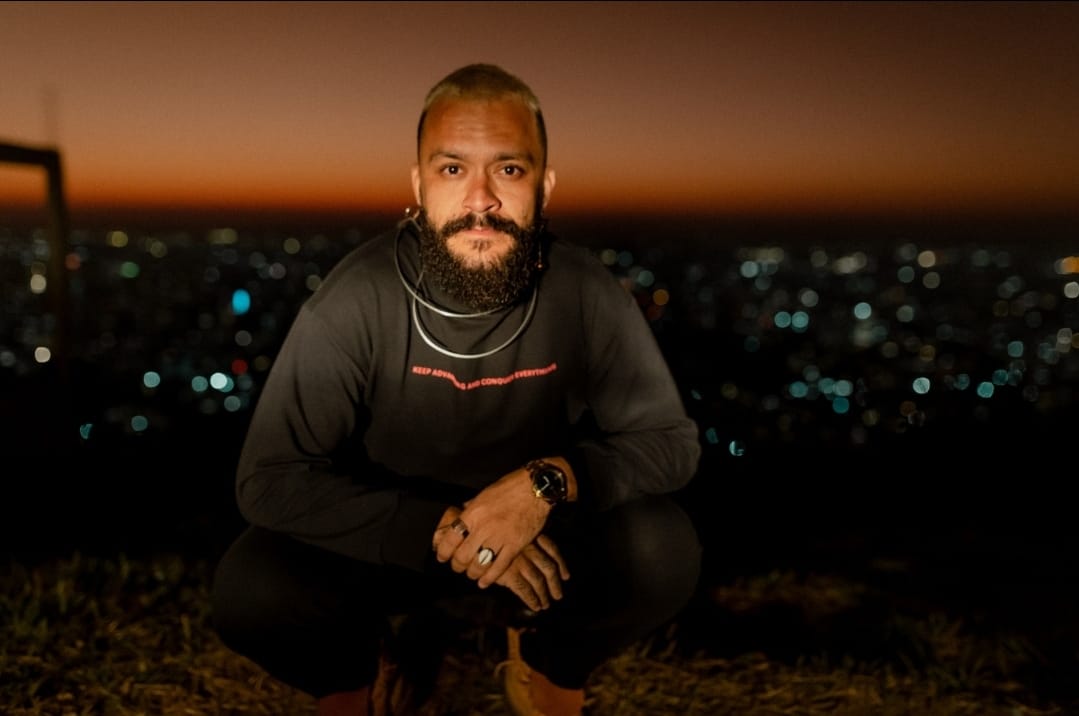 Homem negro, com cabelo raspado e descolorido. Ele usa barba., colares e moletom preto. O homem está agachado segurando as mãos. Ao fundo, desfocado, há um fim de tarde e casas