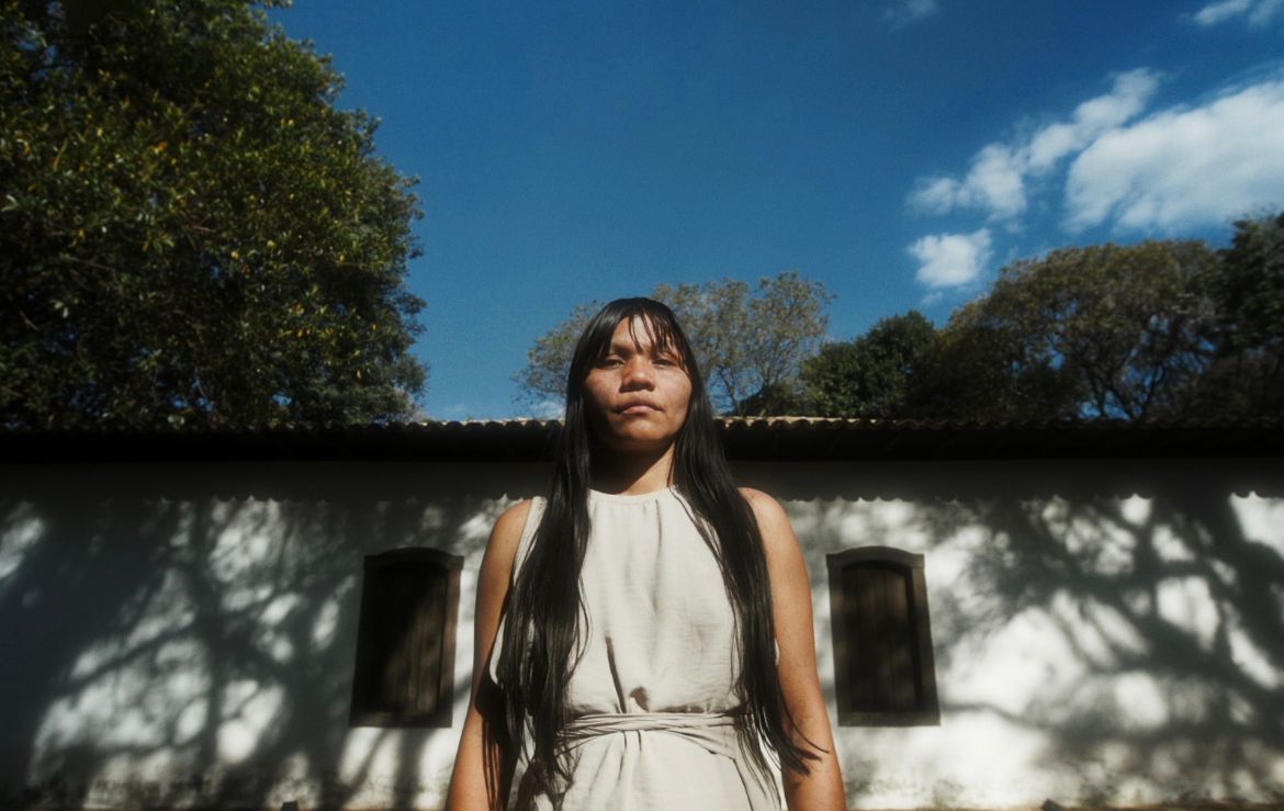 mulher indígena, olhando para câmera. Ela está em pé, com os cabelos longos e soltos, franja e vestido cinza. Ao fundo há uma casa branca, um pedaço do céu azul e árvores na lateral