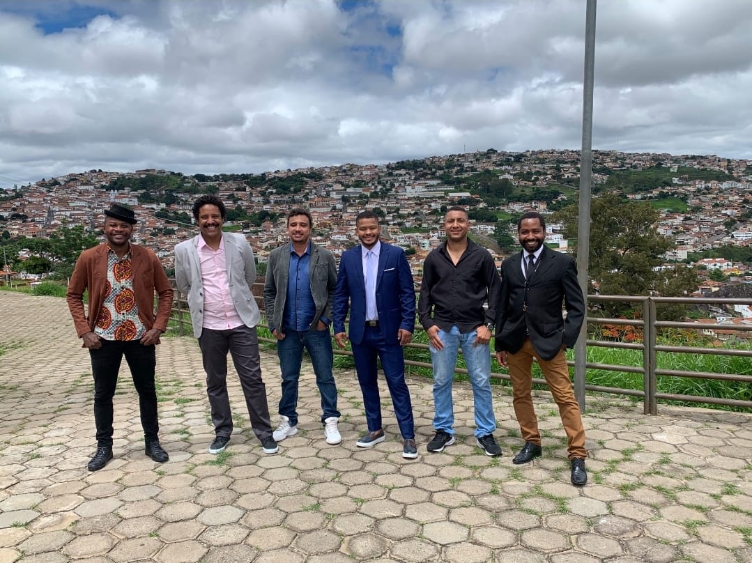 Seis homens negros estão em fila, em pé, olhando para câmera. Eles estão sorrindo. 