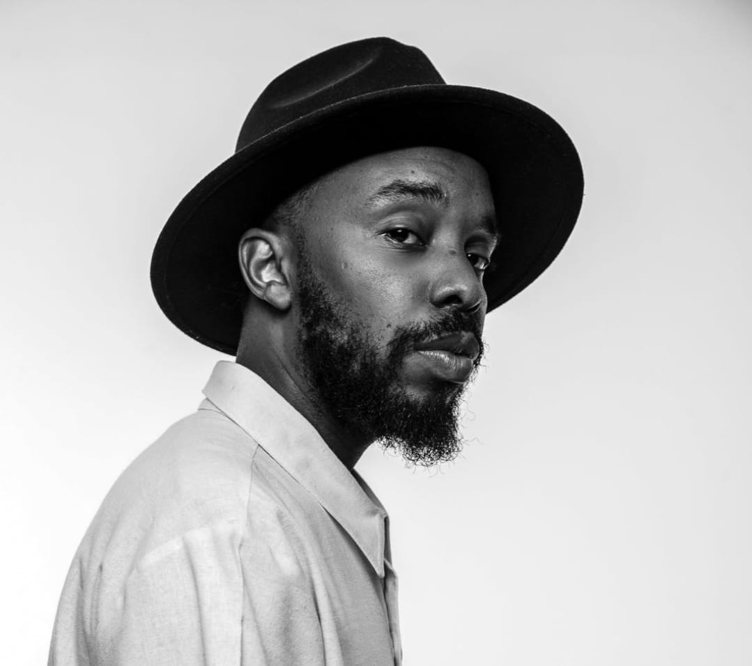 Fotografia em preto e branco de um homem negro, barba e chapéu. Ele está com o corpo próximo a lateral esquerda da foto, virado de lado e olhando para câmera. Ele usa uma camisa social clara
