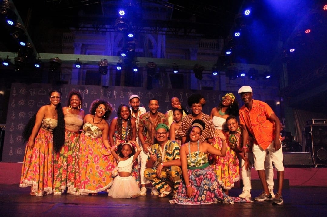 Grupo de pessoas negras posam para foto. Elas estão usando roupas coloridas, as mulheres usam saías rodas. 