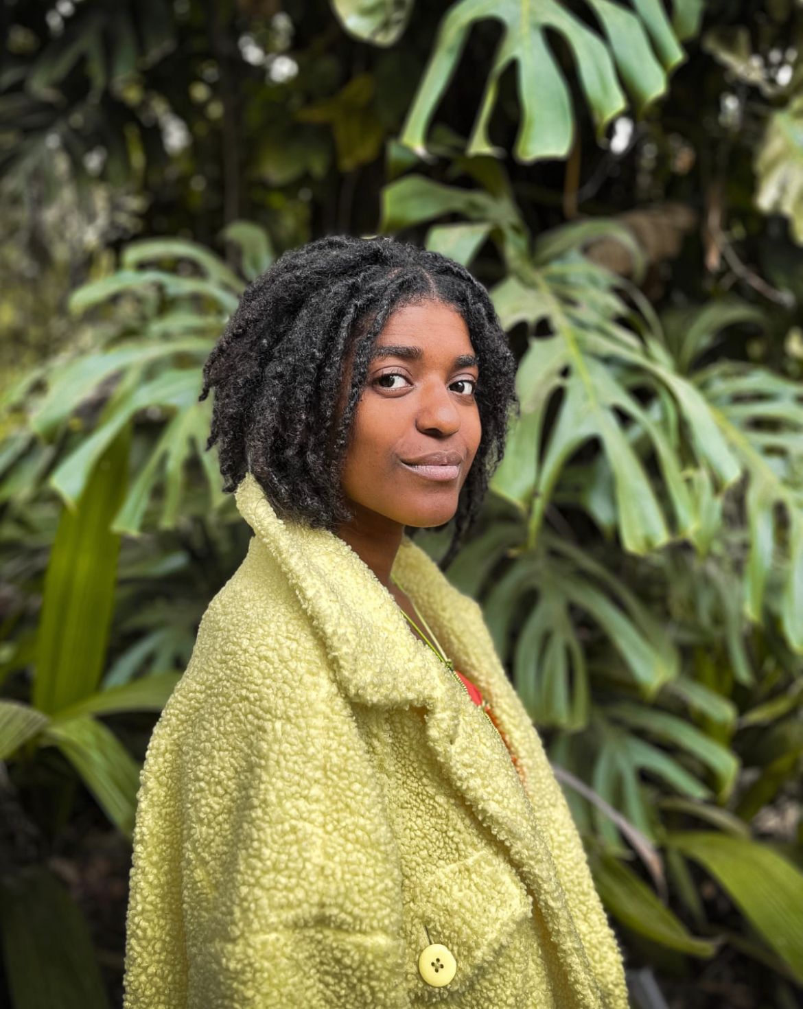 mulher negra olha para câmera. Ela está com o corpo virado para a direita e somente com a cabeça virada para câmera. Ela usa casaco verde e o cabelo curto de dreads. Ao fundo há folhas verdes.