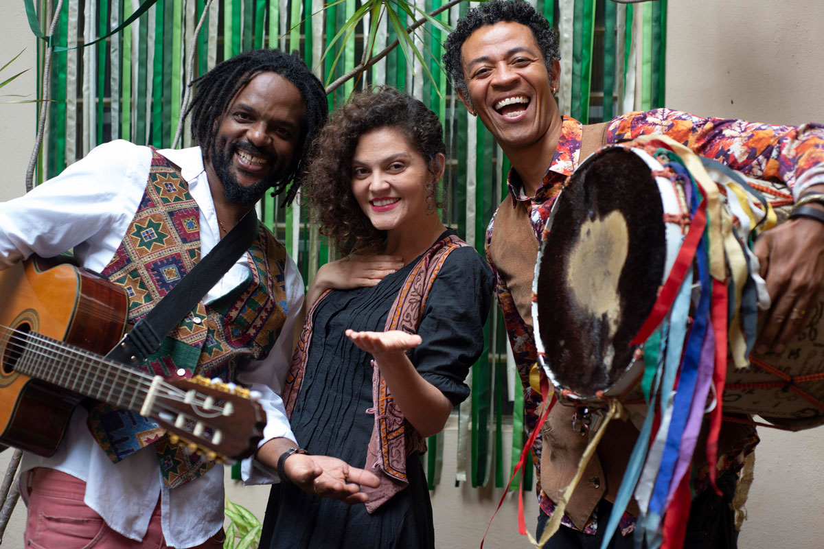 Na foto estão juntos Tininho Silva, um homem negro sorrindo e segurando um violão, Bárbara Amaral, uma mulher branca sorrindo, e Babu Xavier, um homem negro sorrindo e segurando um tambor enfeitado com fitas coloridas, tendo ao fundo fitas verdes dependuradas em uma parede.