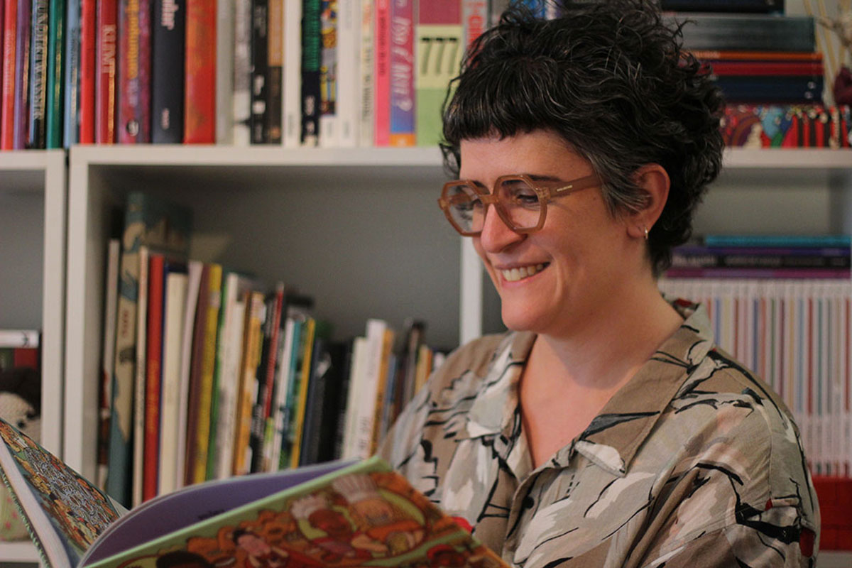 Mulher branca, de cabelos curtos e usando óculos, com um livro em mãos e sorrindo.