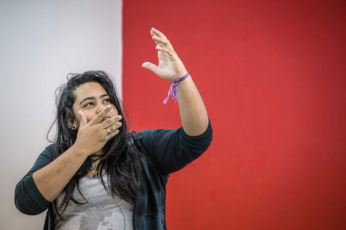 Mulher jovem, de cabelos longos, olhando para o alto. Sua mão direita está próxima ao rosto e a esquerda aponta para o alto. Ao fundo, parede majoritariamente vermelha, com parte de cor cinza.