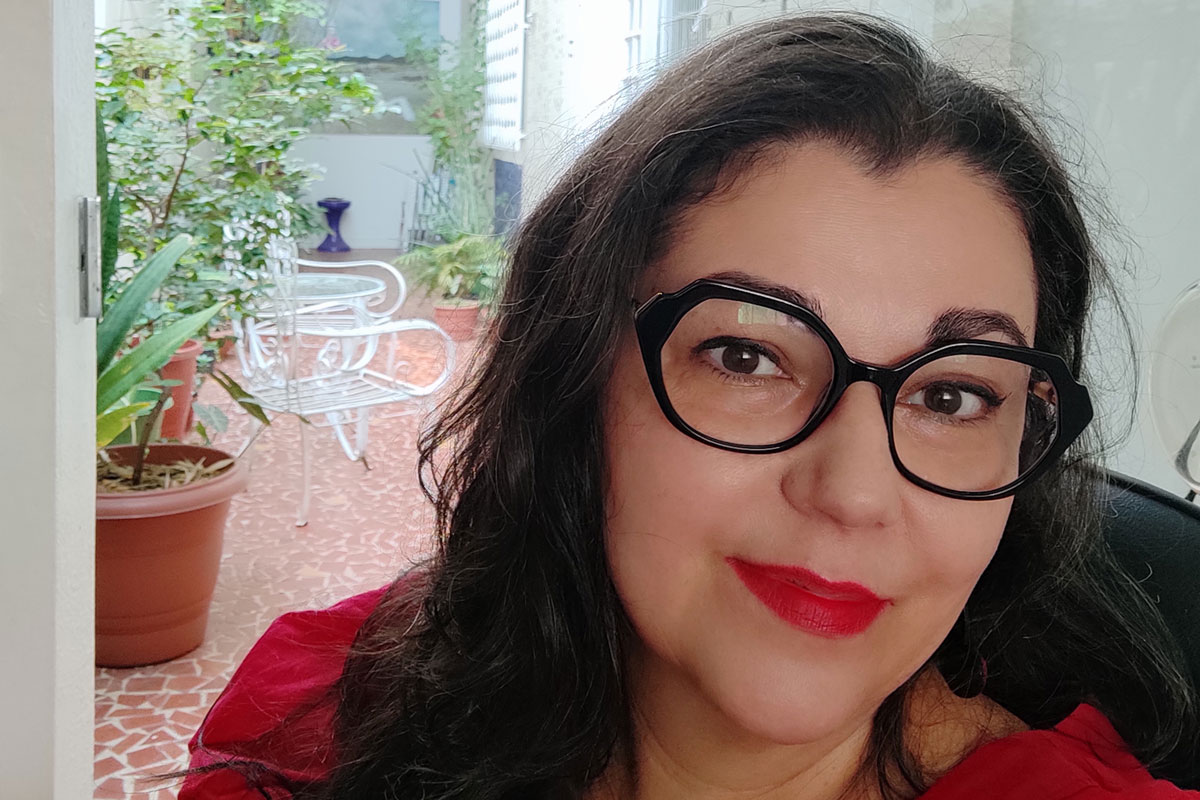 Mulher branca, de cabelos escuros e compridos, usando óculos e sorrindo discretamente.