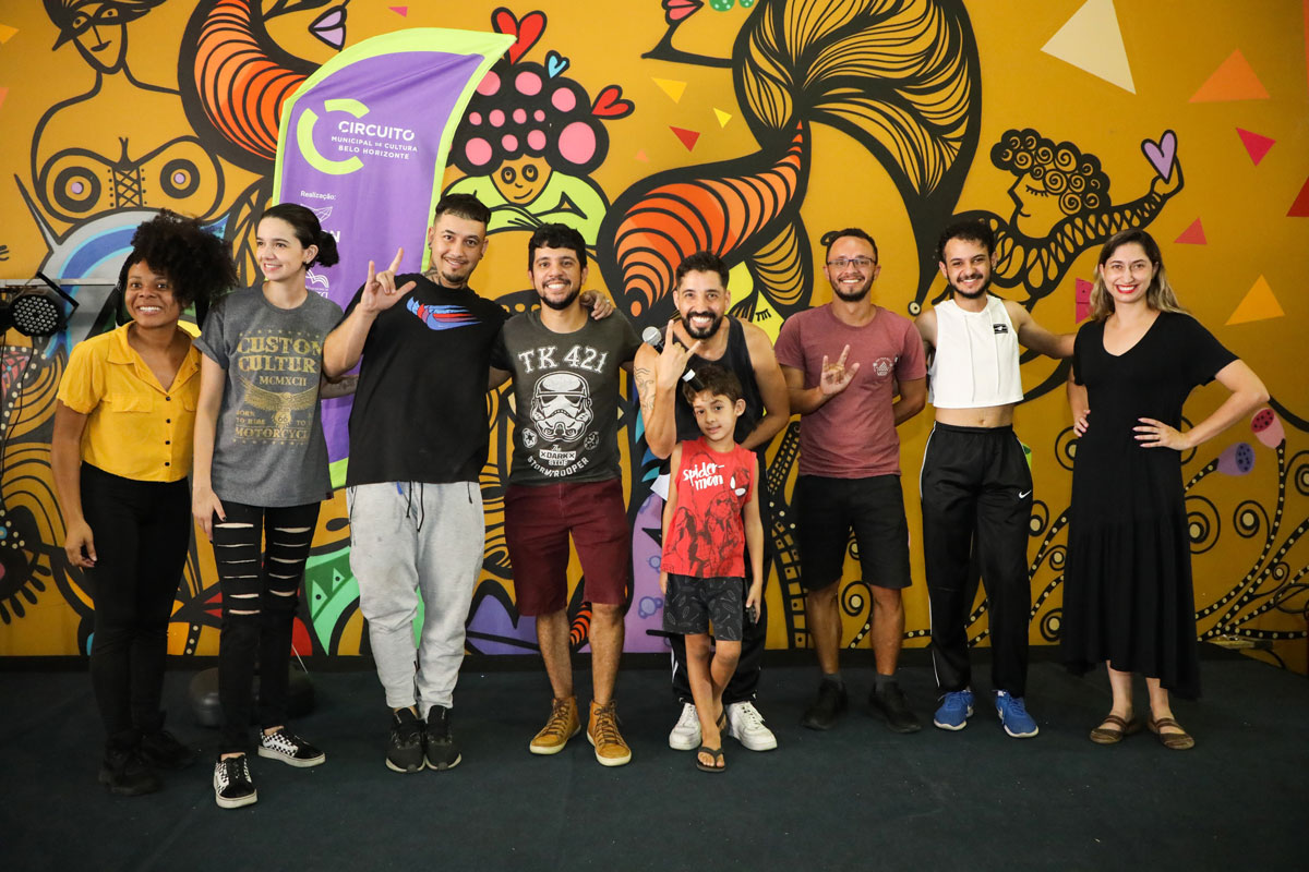 Nove pessoas olham para a frente, sorrindo, tendo ao fundo um mural amarelo com desenhos coloridos.