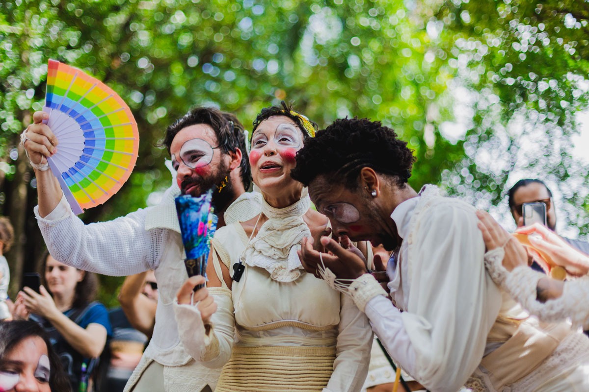Divulgação Três atores vestidos de branco, maquiados, sendo que dois deles estão segurando leques.