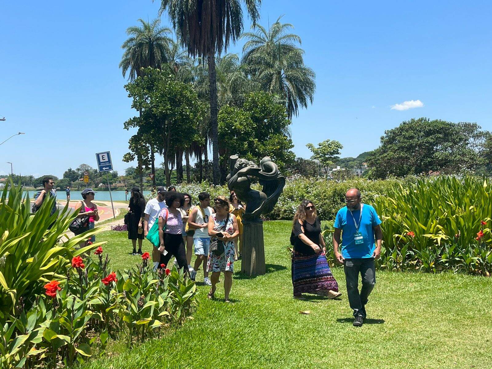 Grupo de pessoas caminha pelos jardins do Museu de Arte da Pampulha. O dia está ensolarado, a grama verde e o céu azul. Há coqueiros ao fundo