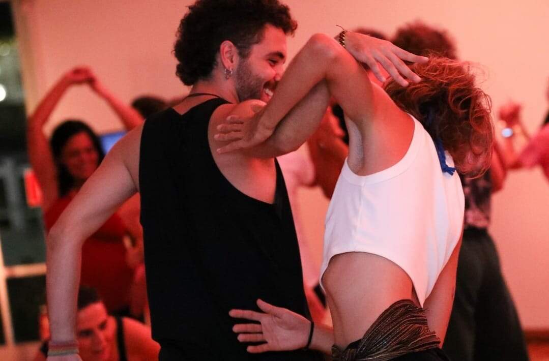 Um homem e uma mulher dançam com os braços entrelaçados. Ele usa camiseta preta e ela camiseta branca