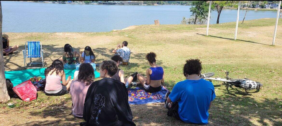 Gupo de pessoas sentadas em um gramado, alguns estão em cima de uma canga. Ao fundo é possível ver parte da Lagoa 