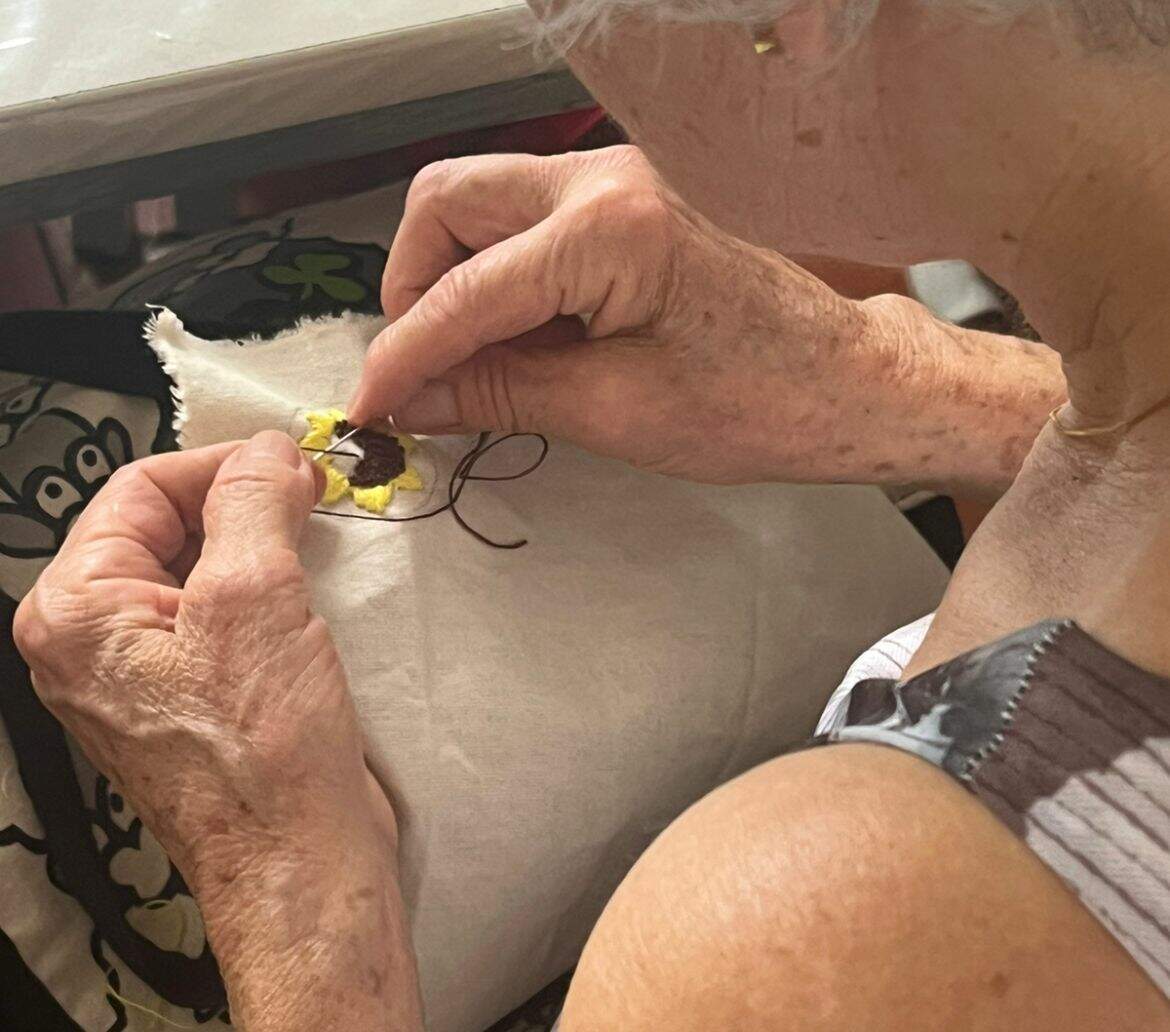 A imagem apresenta as mão de uma mulher branca, idosa, fazendo um bordado. Ela está de costas para câmera