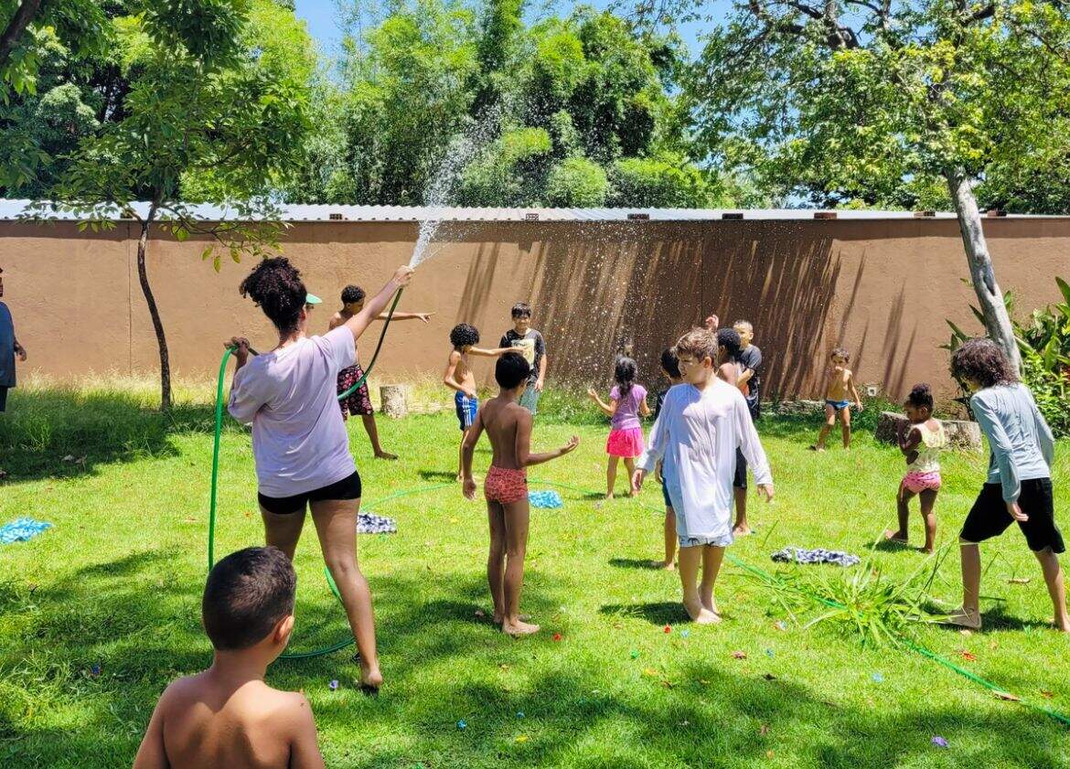 Grupo de crianças tomam banho de mangueira. Uma mulher segura uma mangueira verde, enquanto crianças se divertem no gramado