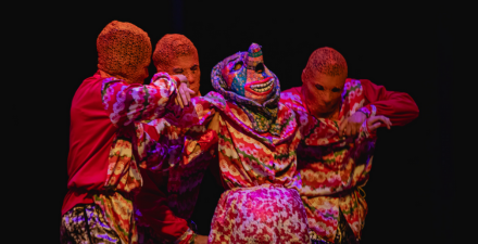 na foto 4 atores usam roupas coloridas e máscaras, 3 atores carregam um personagem que está usando máscara
