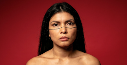 na foto, uma mulher indígena parda de cabelos lisos e pretos. Seu rosto tem uma pintura com uma linha preta marcando pela metade o rosto, abaixo dos olhos em todo o rosto.