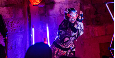 Na imagem uma mulher negra usa uma roupa futurista, um macacão prateado com costuras pretas, capacete e óculos com luzes, e tem luzes roxas ao fundo.