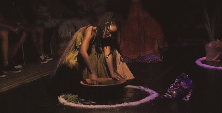 Uma mulher negra com um vestido tomara que caia está agachada no chão usando as mãos para misturar algo em uma vasília de barro. ao redor no chão tem um círculo branco