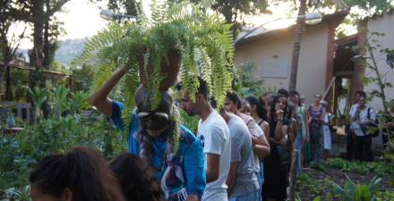 Em fila, pessoas passam pelo quintal de uma casa com horta. Uma personagem com uma máscara e tranças grisalhas leva acima da cabeça um vaso de samambaias.