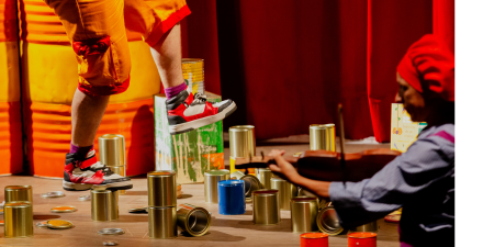 Desimportâncias - Igor Cerqueira Na foto, mostra um enquadramento em que uma pessoa usa roupas coloridas e anda em latas de ferro pelo palco enquanto um homem toca um violino com uma boina azul.