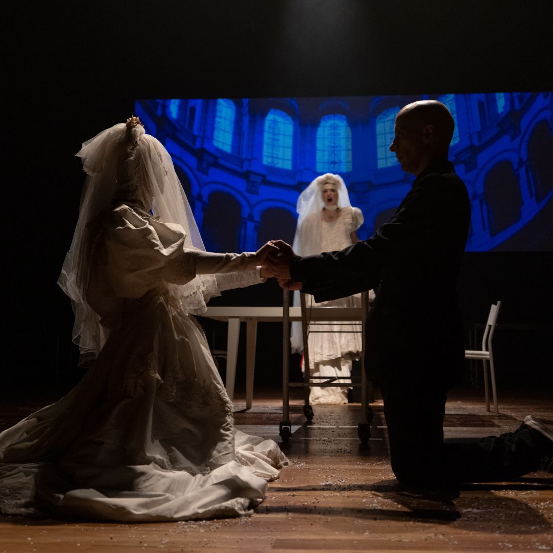 Vestido de Noiva - Foto Netun Lima Na foto, um casal ajoelhado segura as mãos, a noiva com véu, enquanto ao fundo uma outra noiva rege o casamento.