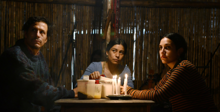 Uma Família chilena sentada à mesa, na luz de velas, se entreolham de forma tensa. Estão sentados à mesa, duas mulheres e um homem.