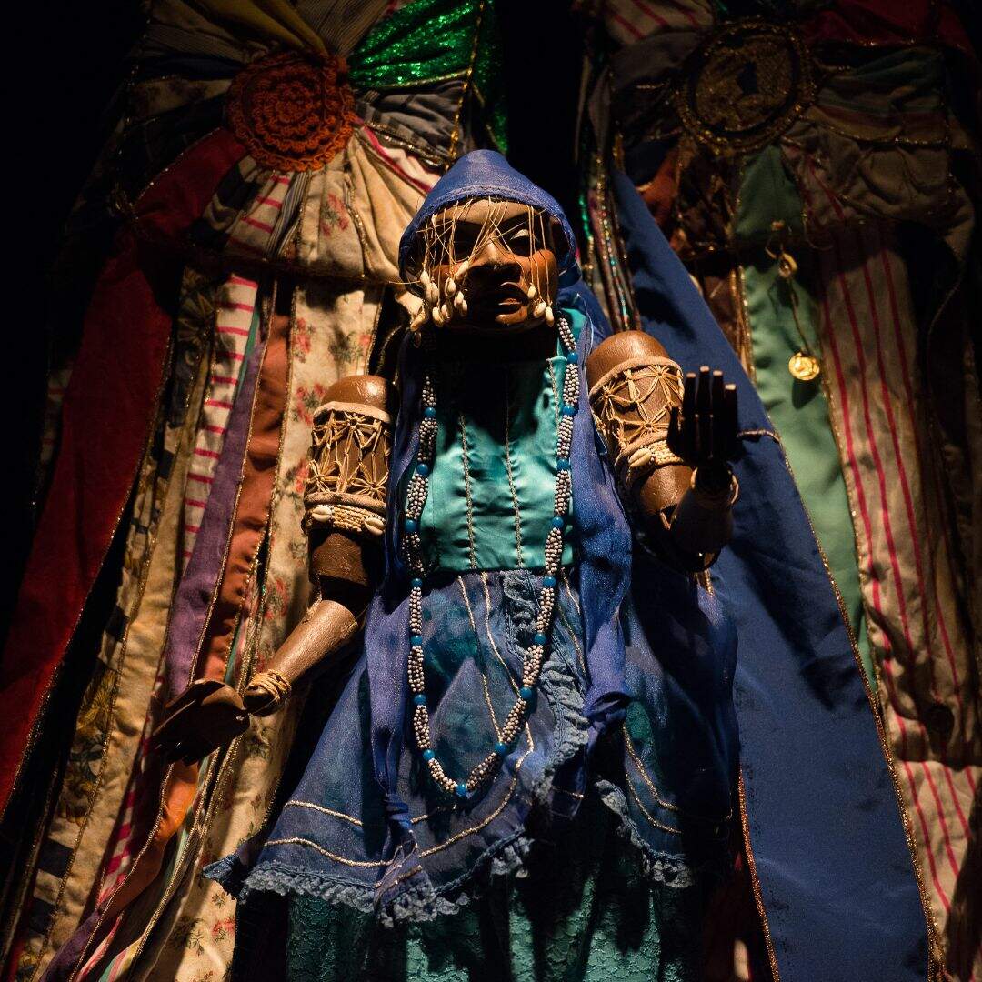 Um boneco habilmente adornado com roupas e colares típicos transmite a aura de um Orixá. Suas vestimentas exibem os símbolos e cores sagradas, refletindo a identidade divina que ele personifica. Com as duas palmas da mão abertas, uma posicionada diante do peito e a outra ao lado da cintura, ele emana uma presença majestosa e poderosa.