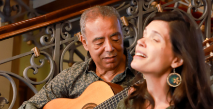 Imagem de um homem e uma mulher em um ambiente interno elegante. O homem, com cabelo grisalho curto, toca um violão e olha atentamente para a mulher, que parece cantar. Ela tem cabelos castanhos longos e usa brincos grandes, em formato circular. O fundo mostra uma escada com um corrimão ornamentado de ferro trabalhado. A atmosfera é intimista, possivelmente de uma apresentação musical ou ensaio.