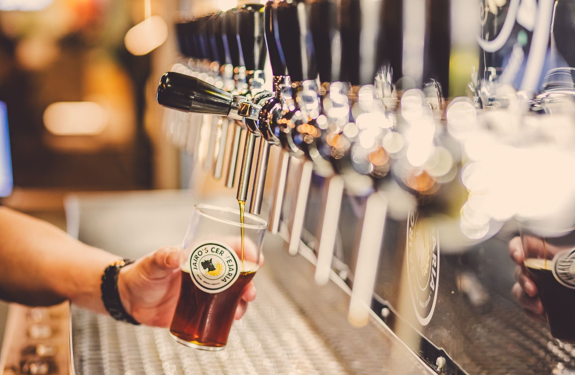 Imagem de varias torneiras de chopp, e uma pessoa enchendo o copo de chopp