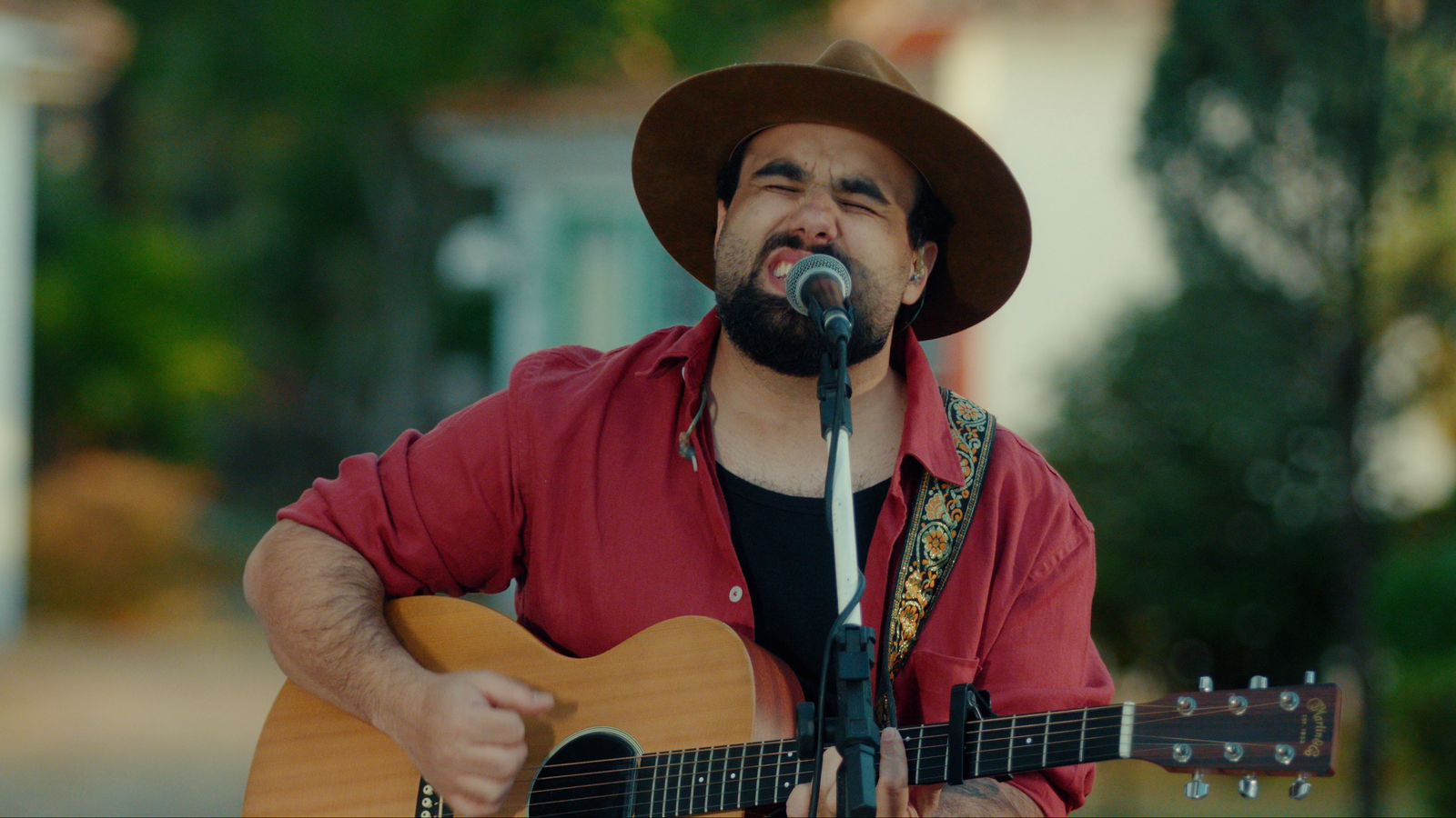 Um homem toca violão e canta ao ar livre. Ele usa chapéu marrom, camisa vermelha e toca diante de um microfone. Atrás dele há casas antigas coloridas e árvores.