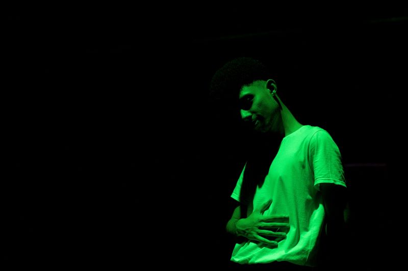 A foto mostra uma pessoa em um ambiente escuro, iluminada por uma luz verde. Ela está usando uma camiseta clara e aparece com uma das mãos sobre o abdômen. O fundo é totalmente preto, destacando apenas o corpo e o rosto sob a iluminação verde.