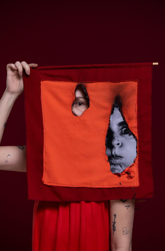 Imagem de uma pessoa segurando em lenço de cor laranja e vermelho, com um buraco no meio e um olho no meio