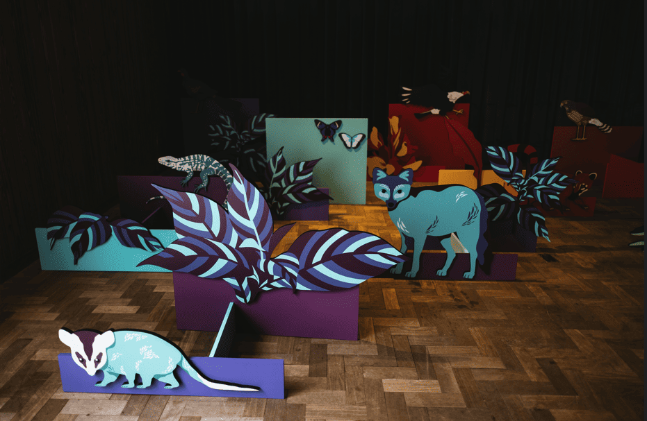 Cenografia em um ambiente interno com figuras de animais da fauna brasileira (como raposa e gambá) e folhagens coloridas espalhadas pelo chão de madeira.