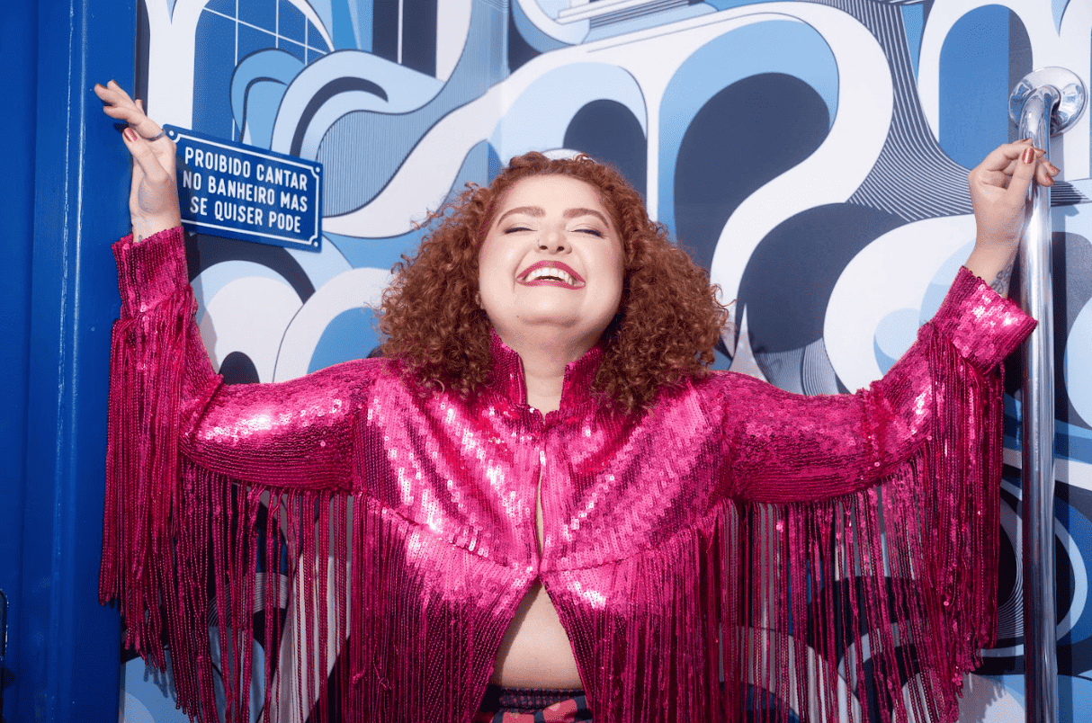 Mulher branca de cabelos cacheados e ruivos sorri com a cabeça para trás, de braços abertos, vestindo uma jaqueta rosa de paetês com franjas. Ao fundo, uma parede com estampa abstrata azul e branca exibe uma placa com os dizeres: "Proibido cantar no banheiro mas se quiser pode".
