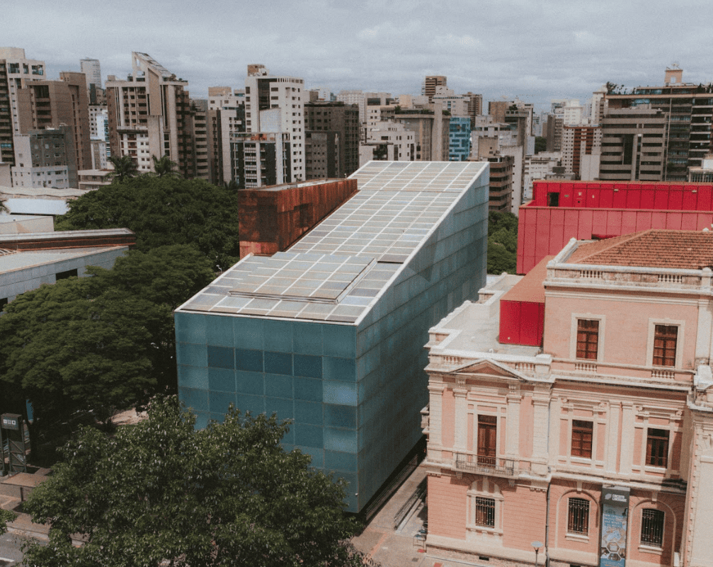 Fotografia aérea de um prédio moderno de formato inclinado com fachada azul e teto coberto por painéis solares. Ele está situado entre edifícios históricos de arquitetura clássica e uma área arborizada, com o horizonte da cidade ao fundo.