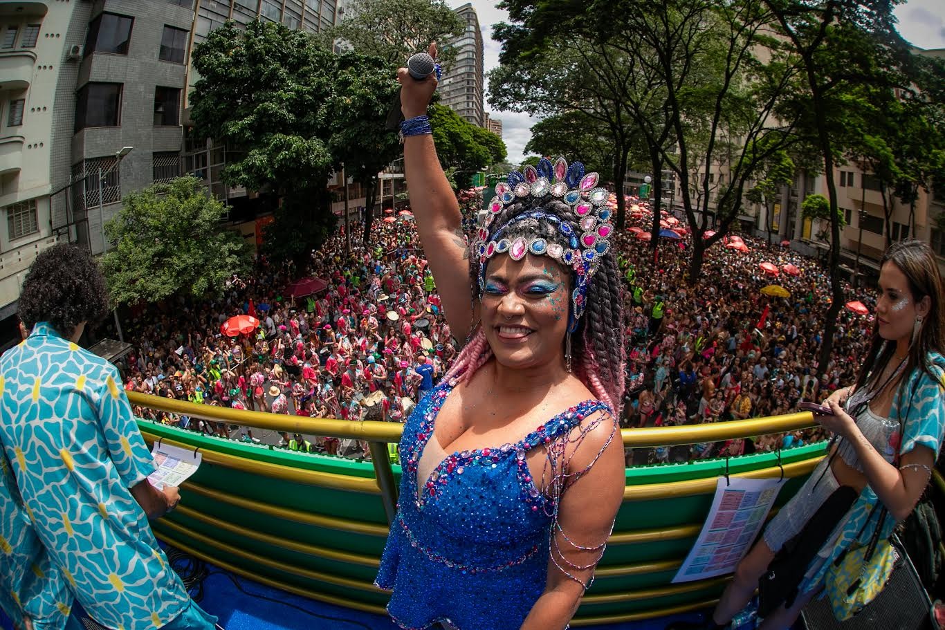 Três pessoas com fantasia de carnaval em cima do trio elétrico. Multidão ao fundo. Ensaio de Carnaval do Bloco Quando Come se Lambuza 2026