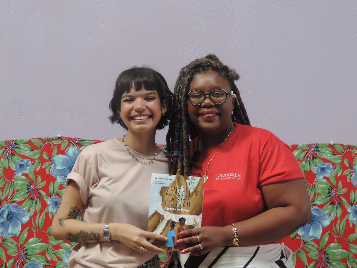 Duas mulheres ao centro segurando o livro "Zenzele e Aziza: através do tempo". Mulher branca à esquerda e mulher preta à direita.