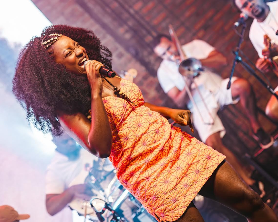 Mulher negra, de cabelos crespos volumosos, está em pé no palco cantando e sorrindo, segurando um microfone com a mão direita. Ela veste um vestido curto laranja com estampa discreta. O ambiente tem iluminação colorida em tons de azul, rosa e verde, e ao fundo há instrumentos musicais e equipamentos de som.