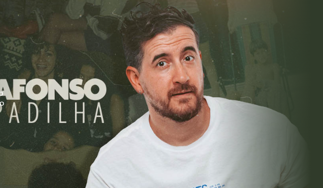 A imagem mostra o comediante brasileiro Afonso Padilha em primeiro plano, centralizado à direita. Ele é um homem branco de cabelos castanhos curtos e levemente bagunçados, barba aparada e usa uma camiseta branca simples. Ele olha para a câmera com uma expressão de dúvida ou questionamento.