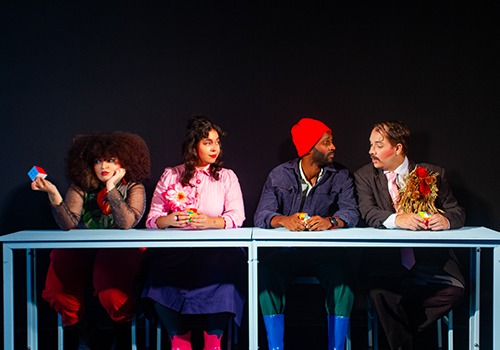 Quatro atores sentados atrás de uma mesa, imagem com fundo preto. Duas mulheres à esquerda e dois homens à direita. As mulheres vestem blusa longa e saia, os homens usam ternos e acessórios coloridos. 