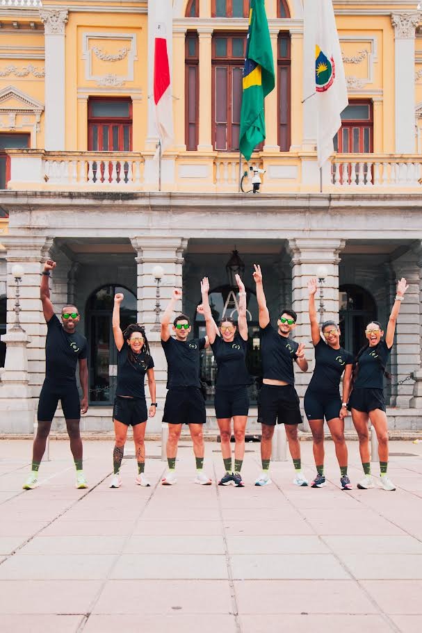 Sete atletas corredores em frente à Praça Rui Barbosa, há três homens e quatro mulheres. Eles vestem blusa e bermuda pretas e erguem um braço em comemoração.
