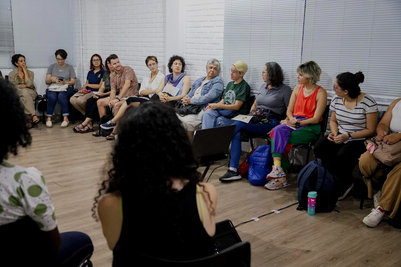 Pessoas sentadas em cadeiras em formato de roda de conversa. Ambiente fechado com paredes branca e cinza e chão marrom.