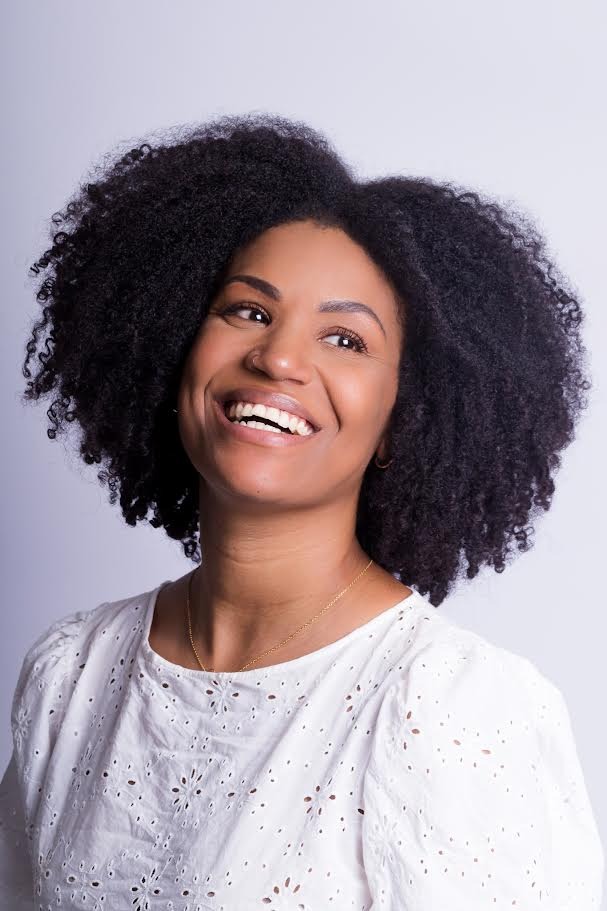 Artista Vavá Fernandes. Mulher preta sorrindo, veste blusa branca. Foto colorida sob fundo claro.