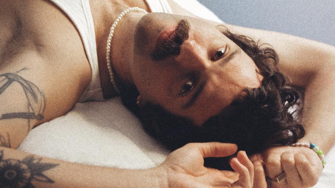 Artista Rafa Castro deitado com os braços próximos à cabeça enquanto encara a câmera com semblante sério. Veste camisa regata branca.